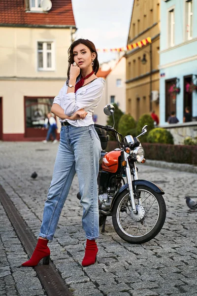 Jovem mulher bonita em vermelho tecido de malha calcanhar calcanhar calça calça jeans azul e casaco de pelúcia posando perto de uma motocicleta em uma rua — Fotografia de Stock