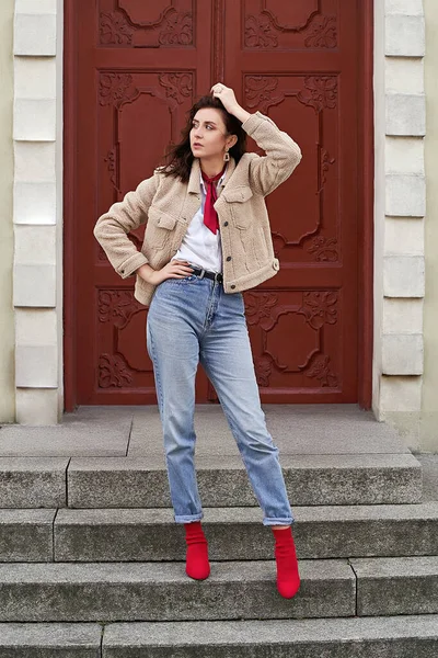Young beautiful woman in red high heel shoes, blue denim jeans pants and teddy jacket coat posing on old red door background in European city — Fotografia de Stock