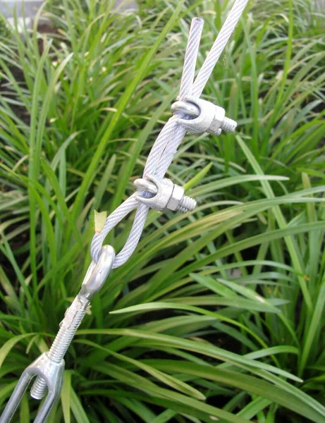 Steel turnbuckle holding hook eye with fastening and metal cables against green grass.