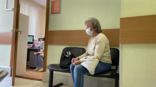 Moskva, Ryssland: En äldre kvinna i ansiktsmask väntar i en sal på kliniken för kvinnor. Kvinnlig klinisk forskning. Åldrad nervös kvinna väntar på läkarbesök — Stockvideo