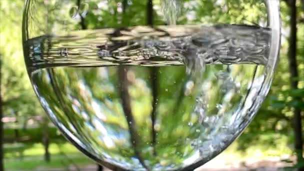 A közeli víz buborékol egy pohárban, vizet öntve egy pohár zöld fák hátterébe egy nyári napon, tiszta vizet öntve lassított felvételen. 4k 38402160. Egészséges életmód koncepció. Buborékáram — Stock videók