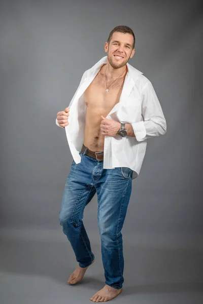 Portrait of a dancing young barefoot man dressed in a white unbuttoned shirt and blue jeans on a gray background in a studio room.