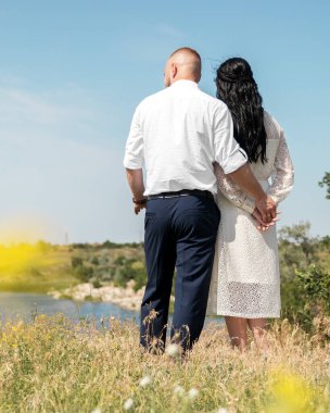 Genç ve mutlu düğün çifti çayırda kameralara sarılıyor ve güneşli yaz gününde ufuk çizgisine bakıyor..