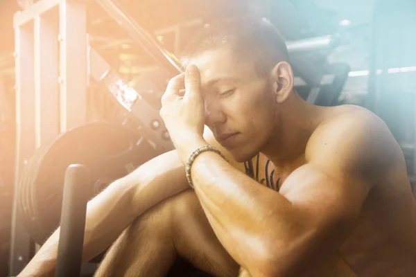 Young athlete sitting on gym equipment with hands on forehead an — Stock Photo, Image