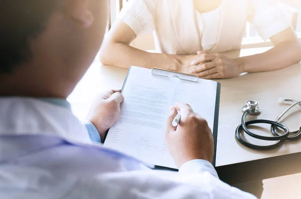 Professional medical doctor in white uniform gown coat interview counseling female patient: Physician writing on patient chart while consultation: Hospital/ clinic healthcare professionalism concept.