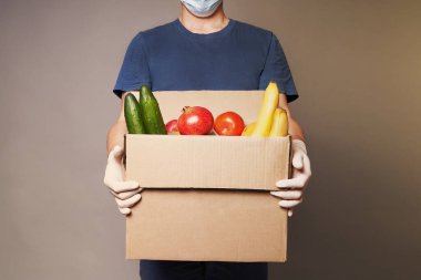 Maske ve lastik eldiven takan beyaz bir erkek kurye bir karton yiyecek kutusu taşıyor.