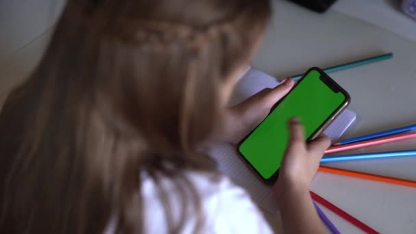 Ein kleines Mädchen sitzt an einem Tisch, das Telefon in der Hand, sie blickt auf die Chroma — Stockvideo