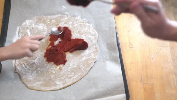 Menina com a mãe cozinhar pizza, esfrega pasta de tomate na massa na forma de um círculo — Vídeo de Stock