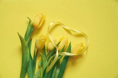 Sarı bir zemin üzerinde beş sarı lale ve kurdeleden yapılmış bir kalp..