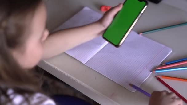 Ένα μικρό κορίτσι κάθεται σε ένα τραπέζι με το τηλέφωνο στα χέρια, κοιτάζει το Chroma — Αρχείο Βίντεο