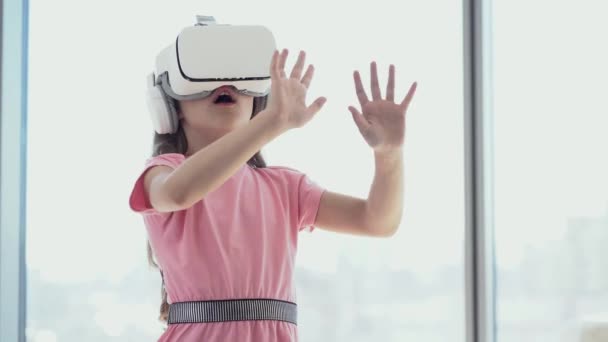 Dívka se baví v bílých brýlích virtuální reality, stojí na pozadí okna a překvapivě se dotýká něčeho rukama — Stock video