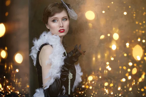 A young beautiful woman in a historical costume of the 20s or 30s of the XX century with a boa and a mouthpiece against the background of lights — Stock Photo, Image