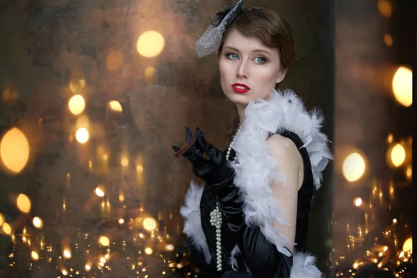 A young beautiful woman in a historical costume of the 20s or 30s of the XX century with a boa and a mouthpiece against the background of lights — Stock Photo, Image