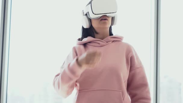 Una ragazza si sta divertendo in occhiali di realtà virtuale, in piedi sullo sfondo di una finestra e giocare a tennis — Video Stock
