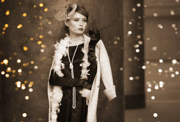 A young beautiful woman in a historical costume of the 20s or 30s of the XX century with a boa and a mouthpiece against the background of lights looks away — Stock Photo, Image