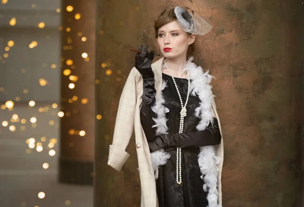 A young beautiful woman in a historical costume of the 20s or 30s of the XX century with a boa and a mouthpiece against the background of lights looks away — Stock Photo, Image