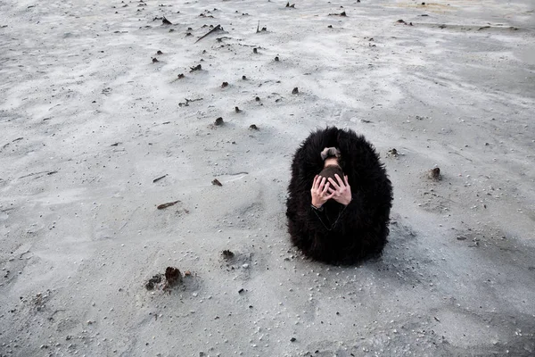Мужчина сидит на белом песке, в черном шубе, с головой в руках. На выжженной кислотой земле. Концепция экологической катастрофы. Карабаш — стоковое фото