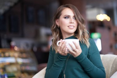 Saç stili ve elinde bir fincan kahve olan güzel bir kadın bir kafede oturuyor. Gülümseyerek yüzünü çeviriyor.