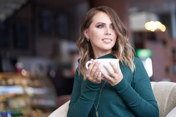 Saç stili ve elinde bir fincan kahve olan güzel bir kadın bir kafede oturuyor. Gülümseyerek yüzünü çeviriyor. — Stok fotoğraf
