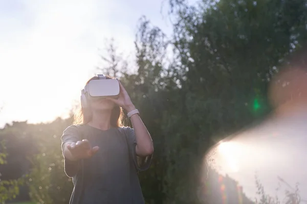 A girl holding virtual reality glasses on her head with her hands. He stretches out one hand to the camera, next to a beam of sunlight.