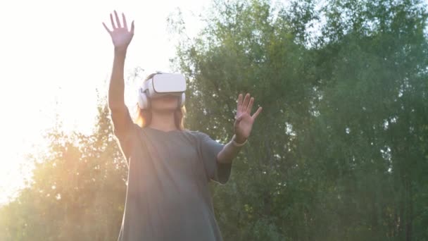 Una ragazza con occhiali di realtà virtuale sulla testa, in piedi sullo sfondo di alberi verdi. Muoveva le mani in aria. Il concetto di futuro. — Video Stock