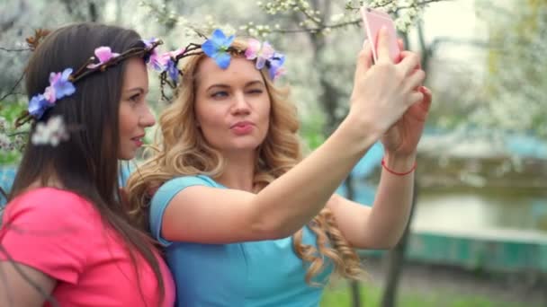 Две девушки в кругах цветов делают селфи в парке весеннего цветения — стоковое видео