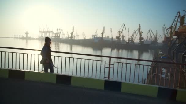 Молодой человек держит скейтборд прогуливаясь вдоль морского порта на рассвете замедленной съемки — стоковое видео