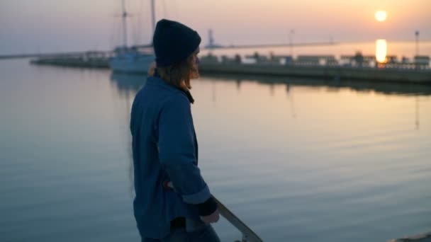 Joven sosteniendo un monopatín caminando a lo largo del puerto marítimo al amanecer — Vídeo de stock