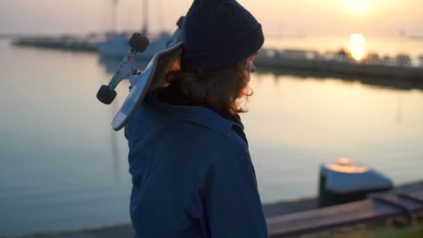 Jonge man met een skateboard wandelen langs de haven aan de zee bij zonsopgang — Stockvideo