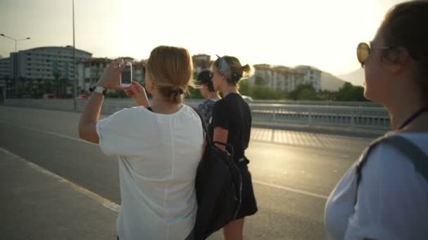 Четыре девушки идут по мосту и фотографируют на смартфоне замедленным движением — стоковое видео