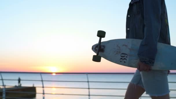 Стрелявший в куклу мужчина несет лонгборд вдоль моря на восходе — стоковое видео