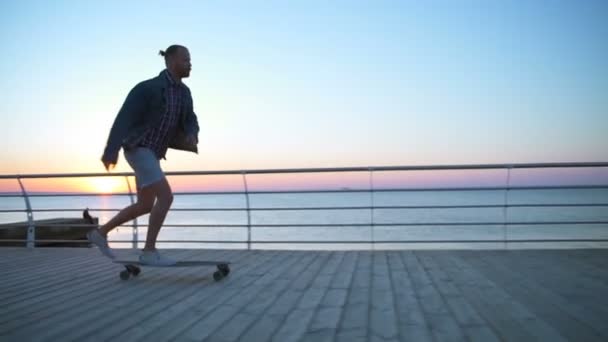 Schöner Mann fährt Longboard entlang des Meeres bei Sonnenaufgang in Zeitlupe — Stockvideo