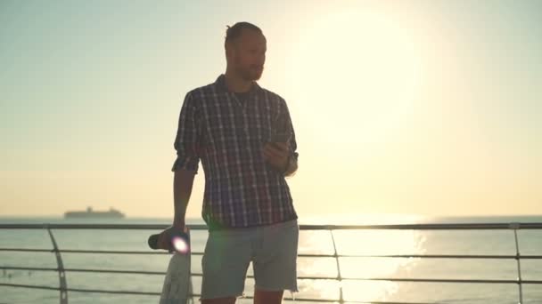 Schöner Skateboarder mit Smartphone mit Meer im Hintergrund in Zeitlupe — Stockvideo