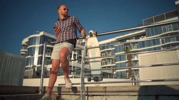 Macho guapo se encuentra cerca de hotel de lujo con teléfono inteligente y longboard — Vídeos de Stock