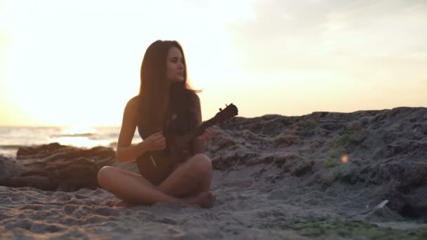 Souriant femelle jouer ukulele dans la lumière du soleil sur la plage au ralenti — Video