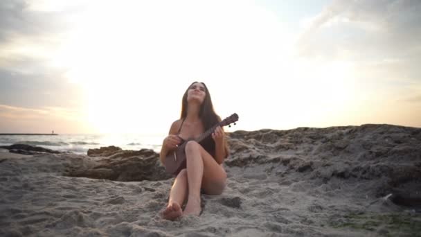 Menina alegre em biquíni joga ukulele na praia câmera lenta — Vídeo de Stock