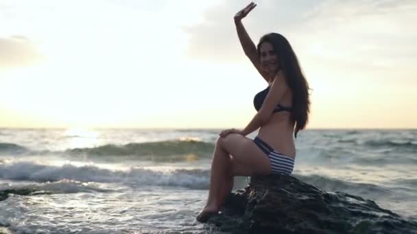 Piękna kobieta siedzi na brzegu morza macha ręką, aby aparat zwolnionym tempie — Wideo stockowe