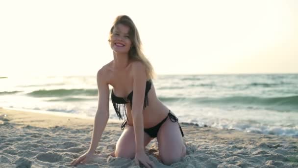 Jeune belle fille en maillot de bain souriant, touchant le sable, assise au bord de la mer — Video