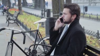 İş adamı bankta oturmuş telefonda konuşuyor.