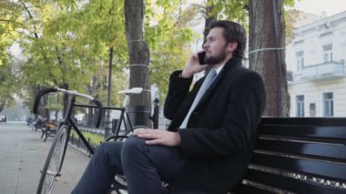 İş adamı bankta oturmuş telefonda konuşuyor.