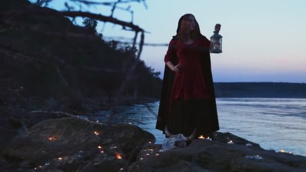 Young female dressed as a witch standing on rocks near the lake — Stock Video