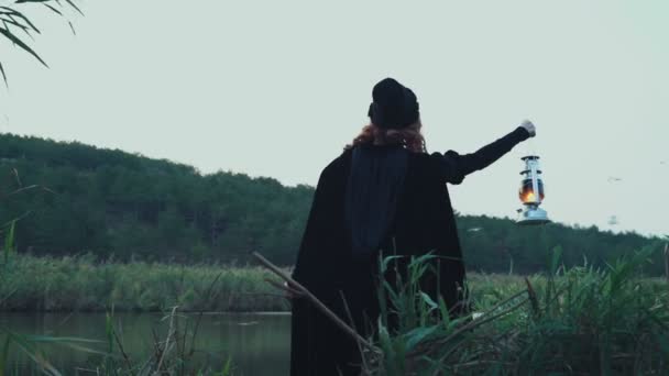 Female wearing cloak holding a lamp standing near the lake — Stock Video