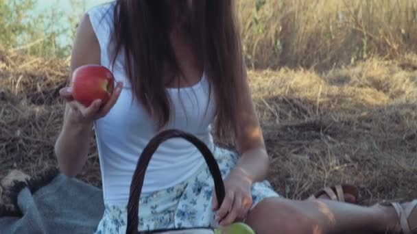 Fille souriante assise sur de l'herbe sèche et prenant une pomme dans un panier de fruits — Video