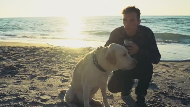 Jeune homme avec son chien sur une plage buvant au ralenti thermocup — Video