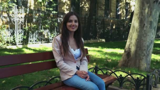 Молодая женщина сидит на скамейке в парке и смотрит в камеру — стоковое видео
