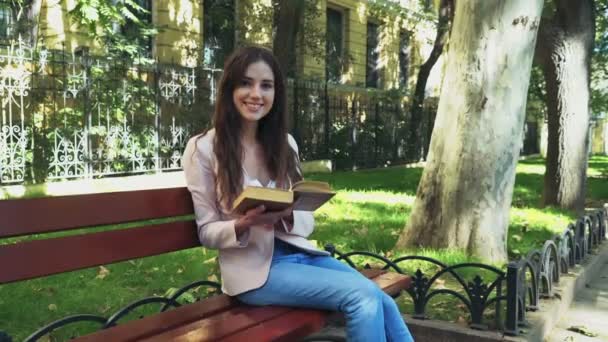 Sorridente estudante do sexo feminino sentado em um banco com um livro e olhando para a câmera — Vídeo de Stock