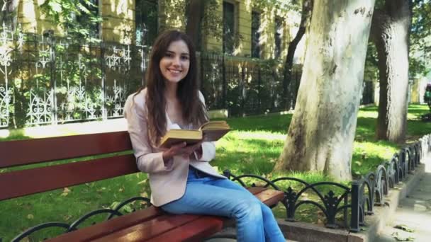 Smiling female student sitting on a bench with a book and looking in camera — Stock Video