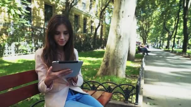 Female student sitting on a bench and using a tablet in park — Stock Video