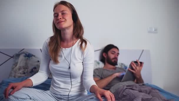 Kvinna lyssna på musik i hörlurar och hane med en tablett i sängen — Stockvideo