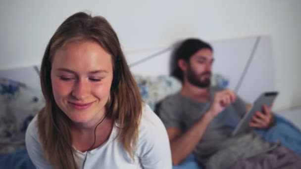 Feminino ouvir música em fones de ouvido e masculino usando um tablet na cama — Vídeo de Stock